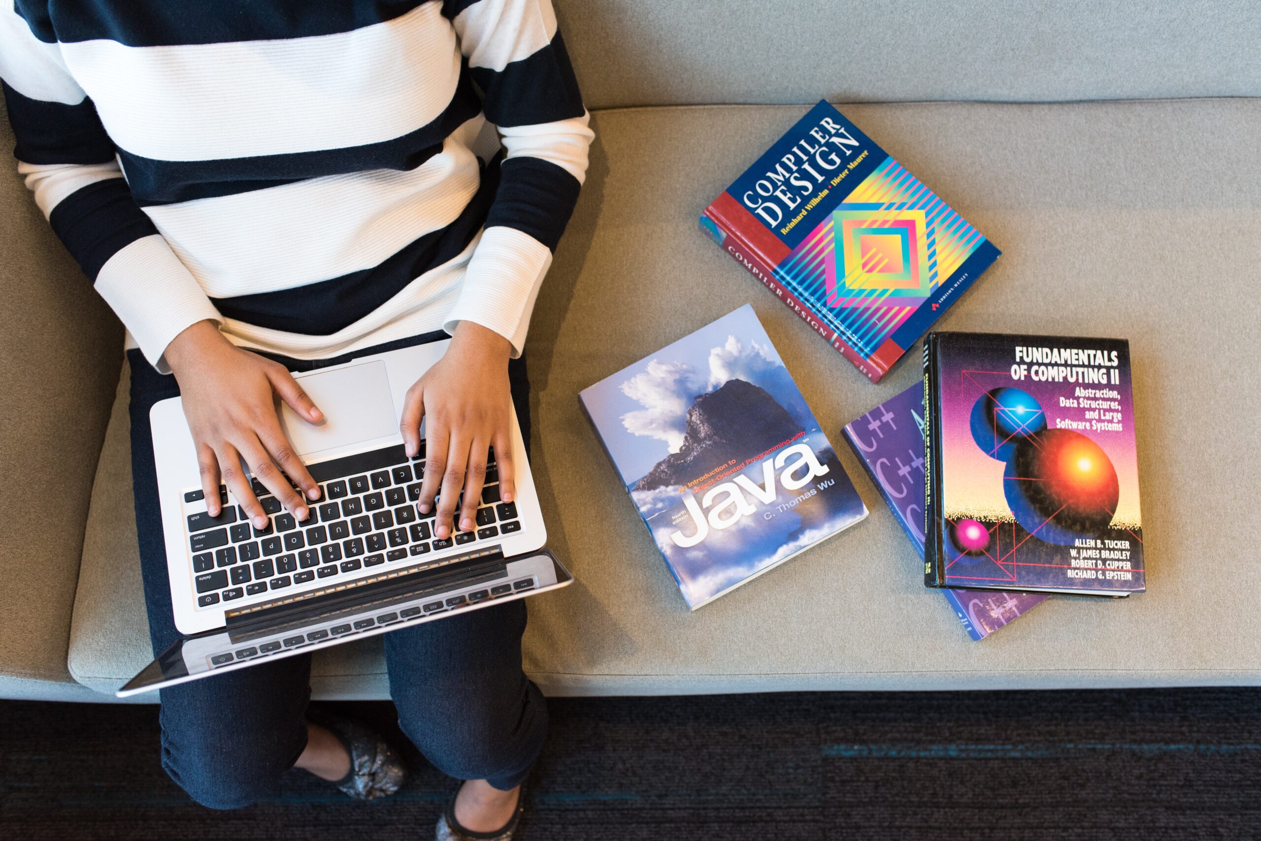 Mulher sentada num sofá com livros e digitando no notebook.