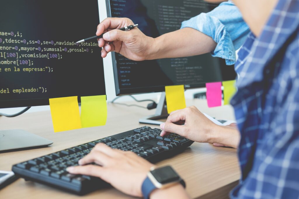 Cover image of the content about Developer platform projects, featuring two individuals engaged in a conversation in front of a computer displaying codes and papers affixed on top, organizing the tasks.