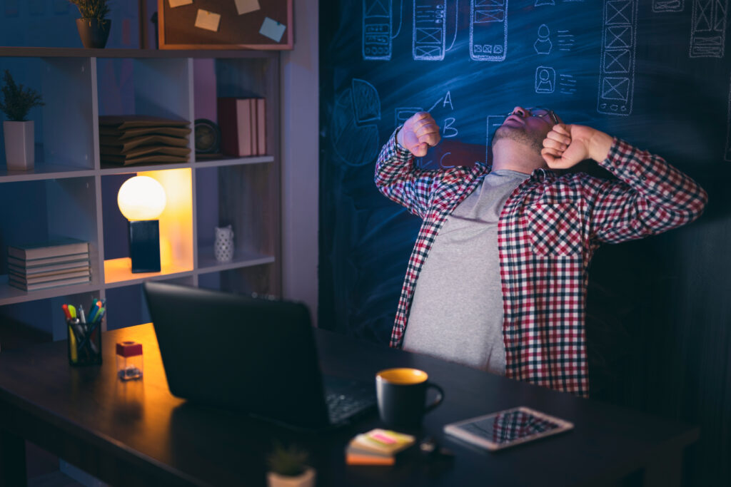 Image cover of the content 'Developer's cognitive overload: How to reduce it on your team?' featuring a web developer stretching while working late. On his desk, there's a black coffee mug, a calculator, and a notebook