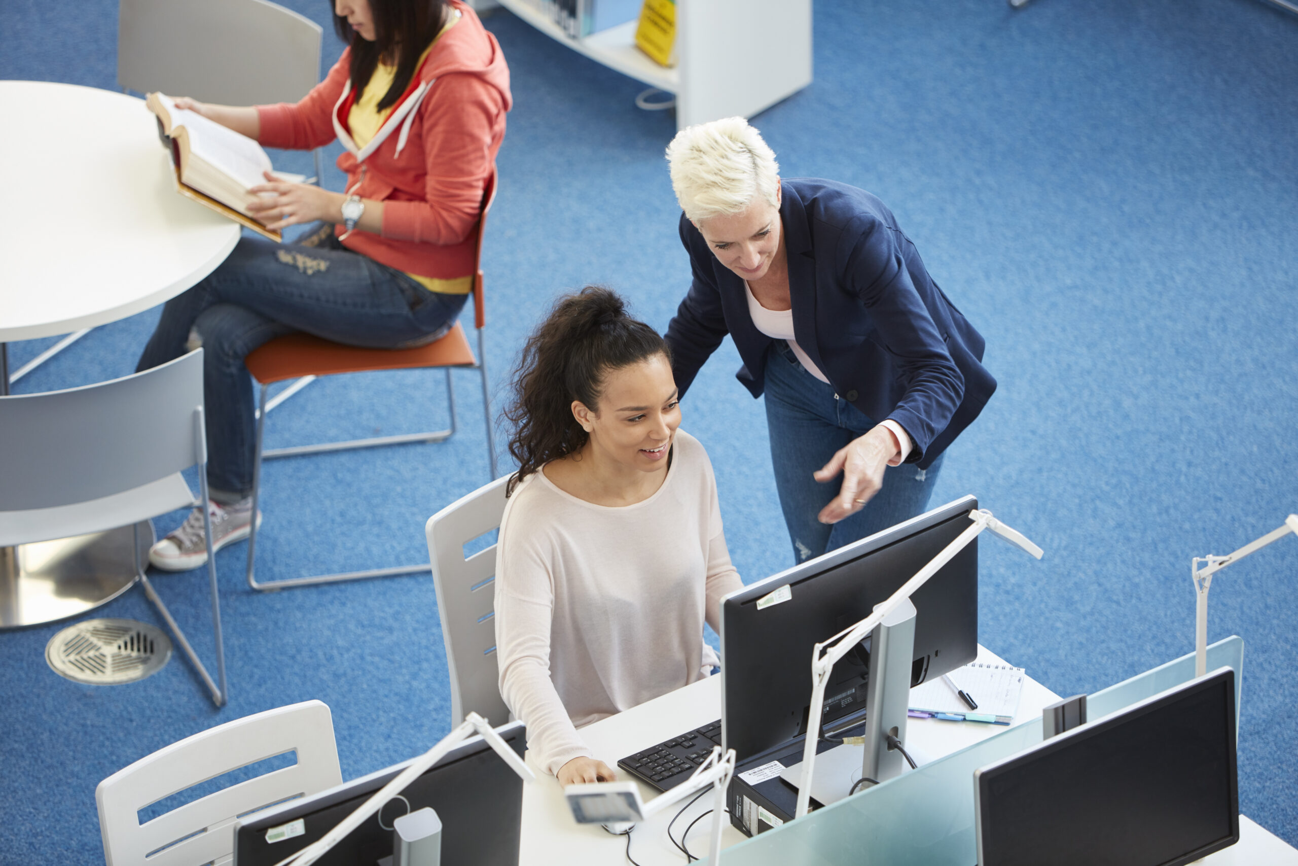 Cover image for the content on knowledge sources, featuring two women in discussion, facing a computer. Additionally, another woman can be seen reading a book at a separate table.