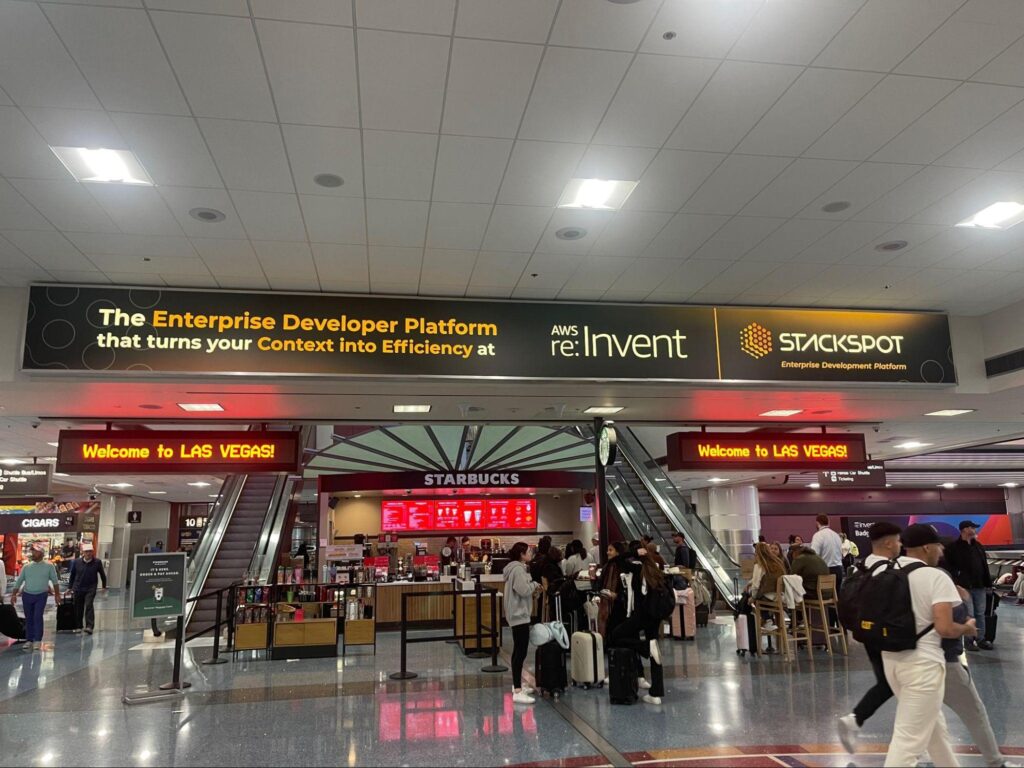Photo of the Las Vegas airport lobby, including a rectangular ad with the phrase The Enterprise Developer Platform that turns your Context into Efficiency at AWS re:Invent 2023 (the latter highlighted). Next to the phrase is the company logo and the words Enterprise Development Platform below. The photo also shows several people passing by and two panels with the greeting Welcome to Las Vegas.