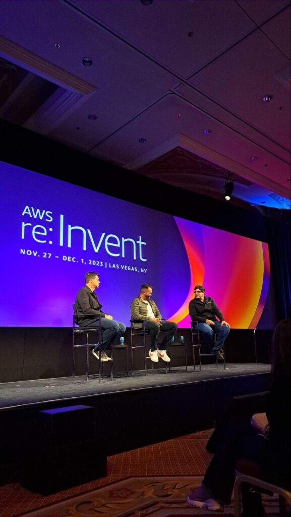 Photo of StackSpot's press conference at AWS re:Invent 2023. Three men are sitting on chairs in front of an audience; the background shows the visual identity of the event in purple, orange, pink and red.