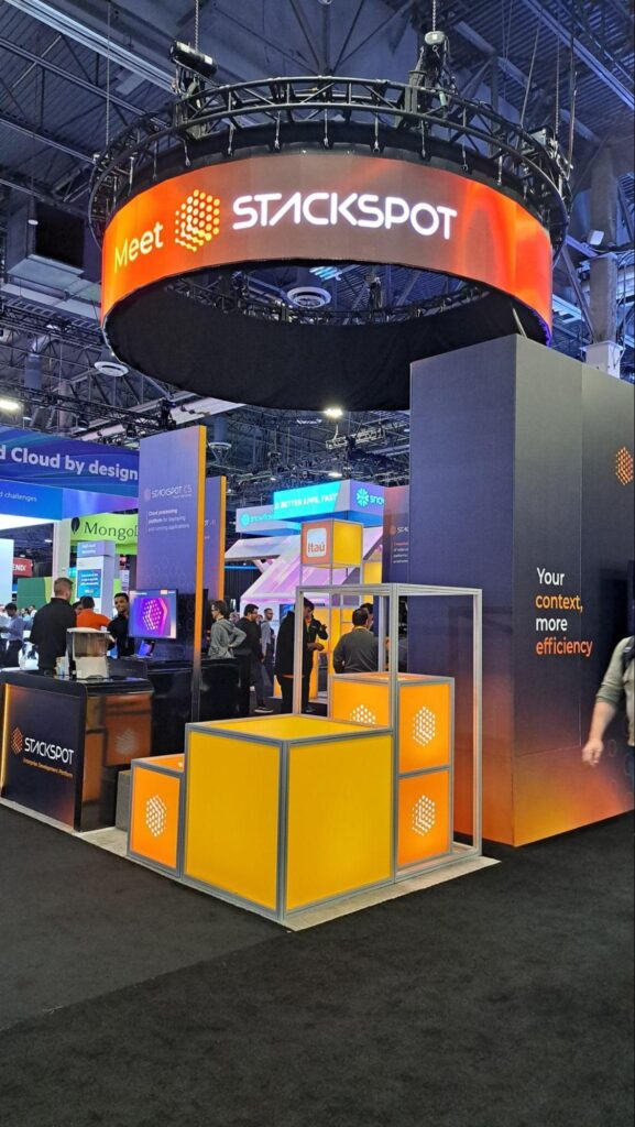 Photo of the StackSpot booth at AWS re:Invent 2023. The LED display on the top has Meet StackSpot written on it; this LED display has a horizontal circular structure, so the message runs in 360º. Below there are large panels with phrases about StackSpot's proposal, such as Your context, more efficiency. In the foreground, orange cubes carry the StackSpot logo.