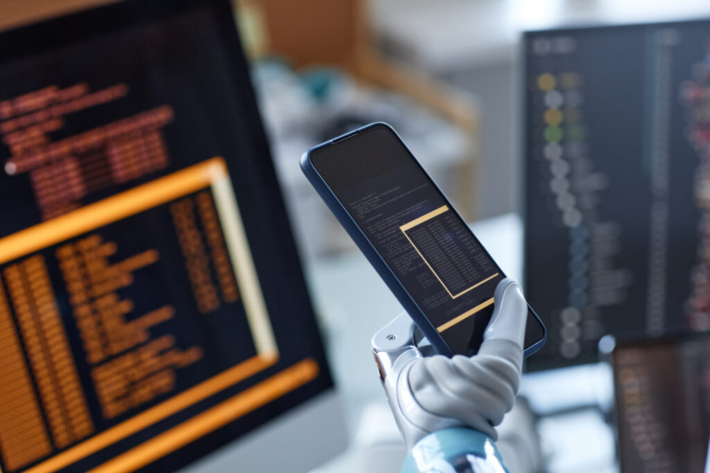 Foto de um celular com códigos aparecendo em sua tela sendo segurado por uma mão biônica. No fundo, estão telas de computador também com códigos.