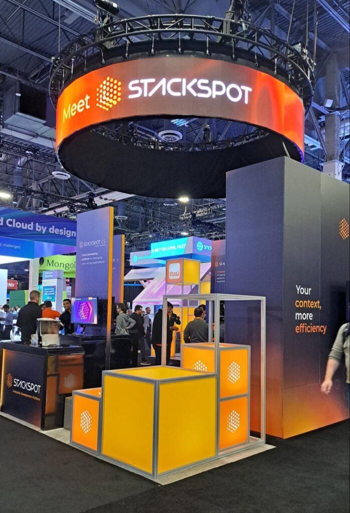 Foto do estande da StackSpot no AWS re:Invent 2023. Logo no alto está um LED com Meet StackSpot escrito nele, esse LED possui a estrutura circular na horizontal, o que faz com que a mensagem seja transmitida em 360º. Abaixo temos grandes painéis com frases acerca da proposta da StackSpot, como Your context, more efficiency. No primeiro plano, temos cubos em laranja com a logo da StackSpot.