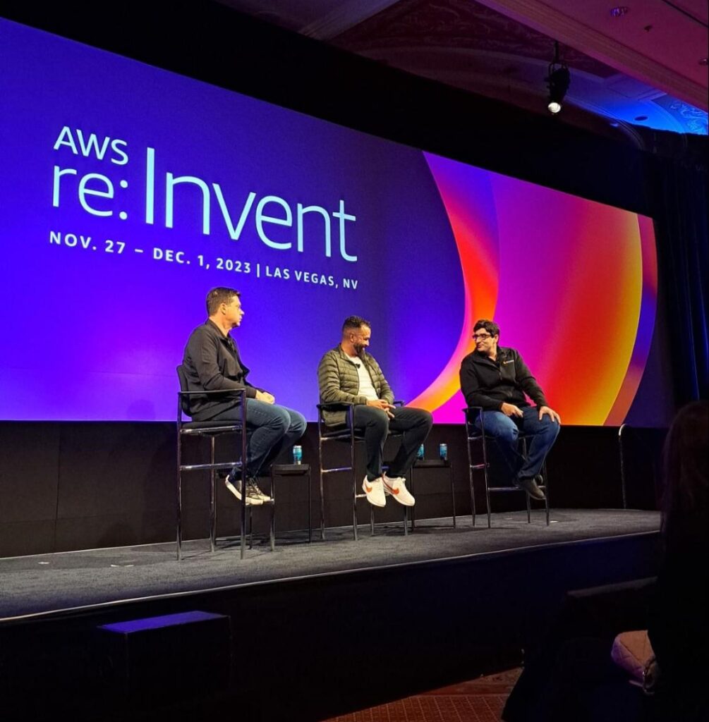 Foto da coletiva de imprensa da StackSpot no AWS re:Invent 2023. Três homens estão sentados em cadeiras em frente à plateia, ao fundo há a identidade visual do evento em tons de roxo, laranja, rosa e vermelho. 