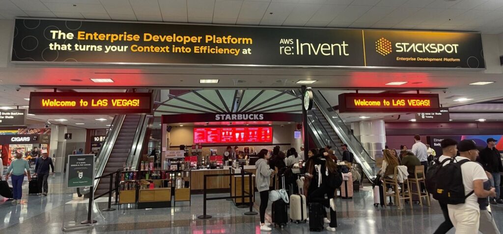 Foto do saguão do aeroporto de Las Vegas, como destaque é possível ver um anúncio retangular com a frase The Enterprise Developer Platform that turns your Context into Efficiency at AWS re:Invent 2023 (este último em destaque). Ao lado da frase está a logo da empresa e StackSpot, abaixo Enterprise Development Platform. Na foto também estão diversas pessoas passando e dois painéis com Welcome to Las Vegas.