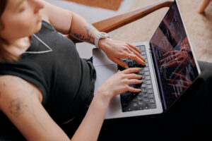 Mulher branca com tatuagens nos braços e uma blusa preta está sentada com o notebook no colo. A foto foi tirada de cima, onde podemos ver as mãos da mulher no teclado e códigos na tela.
