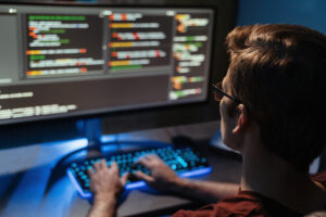 Visão traseira do homem programando e trabalhando em casa até tarde da noite. O homem está em primeiro plano com foco e a tela do computador com códigos está desfocada.