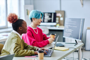 Imagem com duas pessoas jovens em frente ao monitor com códigos na tela. Uma das pessoas é uma mulher negra de cabelo crespo e ao lado um homem de cabelo curto e azul.