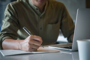 Foto de um homem no plano de fundo de camisa social escrevendo a caneta em um caderno e um notebook ao lado.