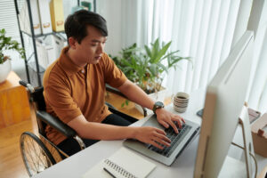 Foto de um homem amarelo em uma cadeira de rodas em frente ao computador com as mãos no teclado.