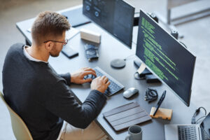 Visão do alto de um desenvolvedor, um homem branco, de cabelos curtos e castanhos, óculos de grau com armação preta e barba, escrevendo código enquanto usa sistemas de computador e dados no escritório